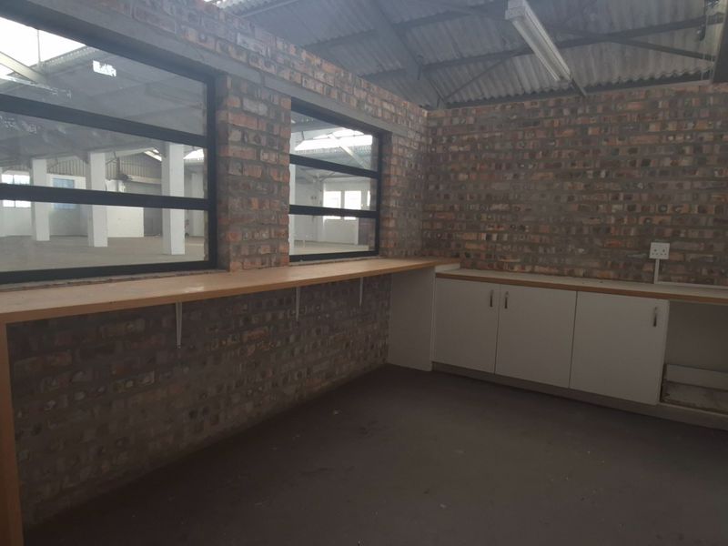 An industrial utility or office space featuring exposed brick walls, built-in cabinetry with laminate countertops, and large metal-framed windows overlooking the warehouse floor.