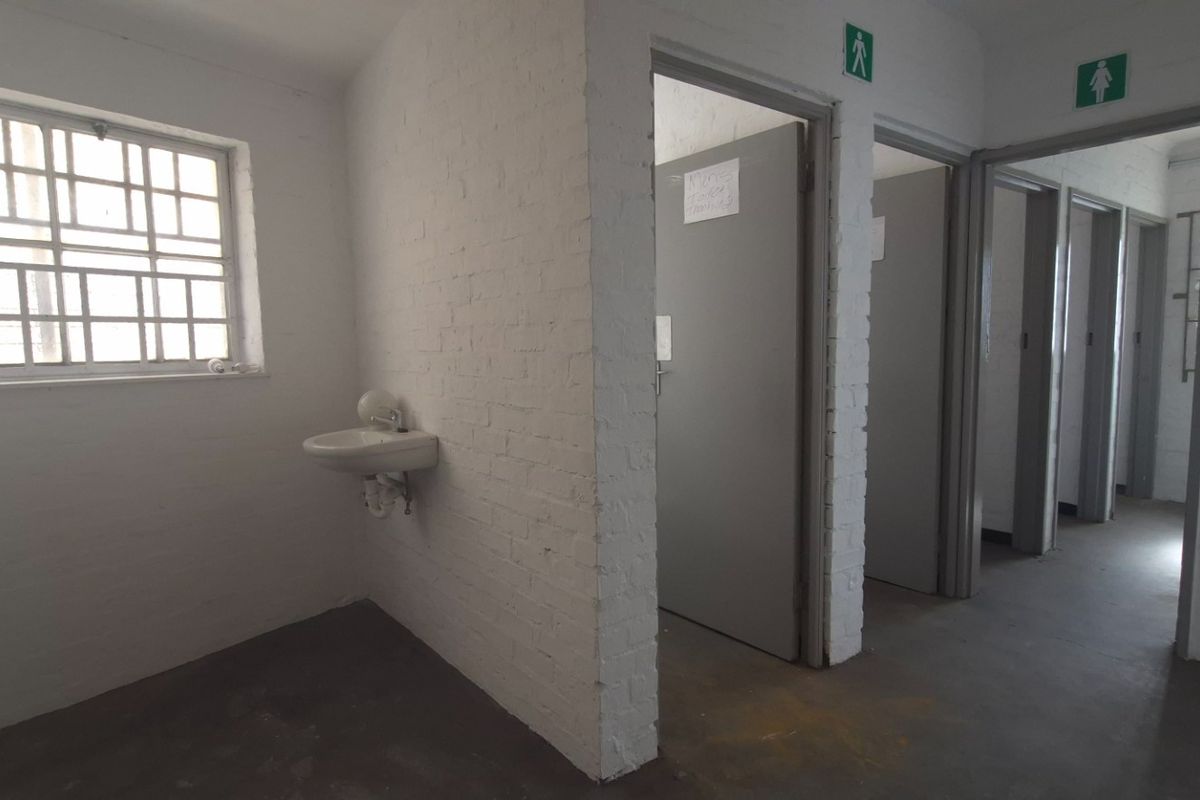 Industrial-style communal bathroom facility featuring multiple cubicles, a wall-mounted basin, and painted brick walls with security-barred windows.