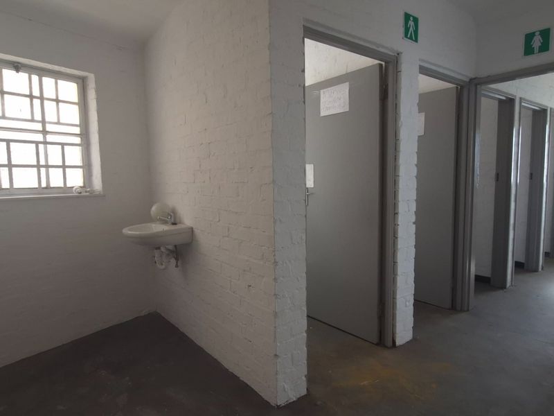 Industrial-style communal bathroom facility featuring multiple cubicles, a wall-mounted basin, and painted brick walls with security-barred windows.
