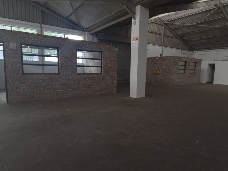 Spacious industrial warehouse interior featuring exposed steel roof trusses, concrete flooring, and partitioned brick office or utility areas with industrial steel-framed windows.