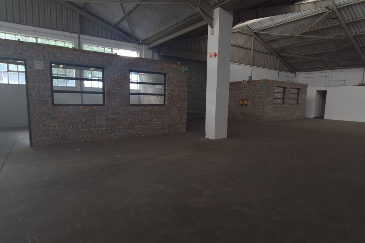 Spacious industrial warehouse interior featuring exposed steel roof trusses, concrete flooring, and partitioned brick office or utility areas with industrial steel-framed windows.