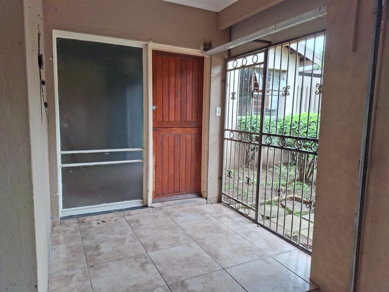 A covered patio area featuring tiled flooring, a wooden entrance door, and a metal security gate overlooking the garden.