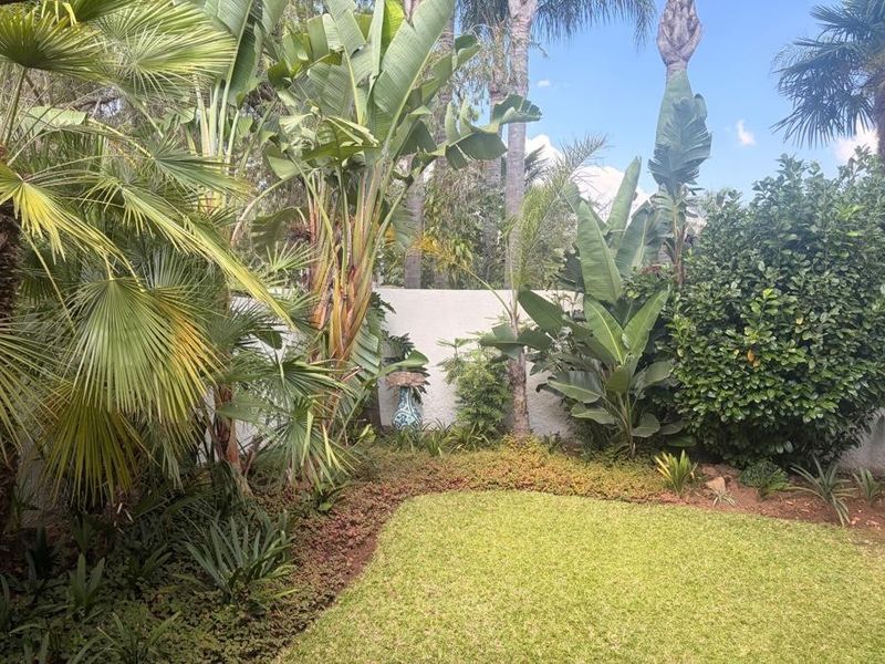 A lush, well-maintained private garden featuring mature tropical plants, palm trees, and a manicured lawn enclosed by a solid boundary wall.