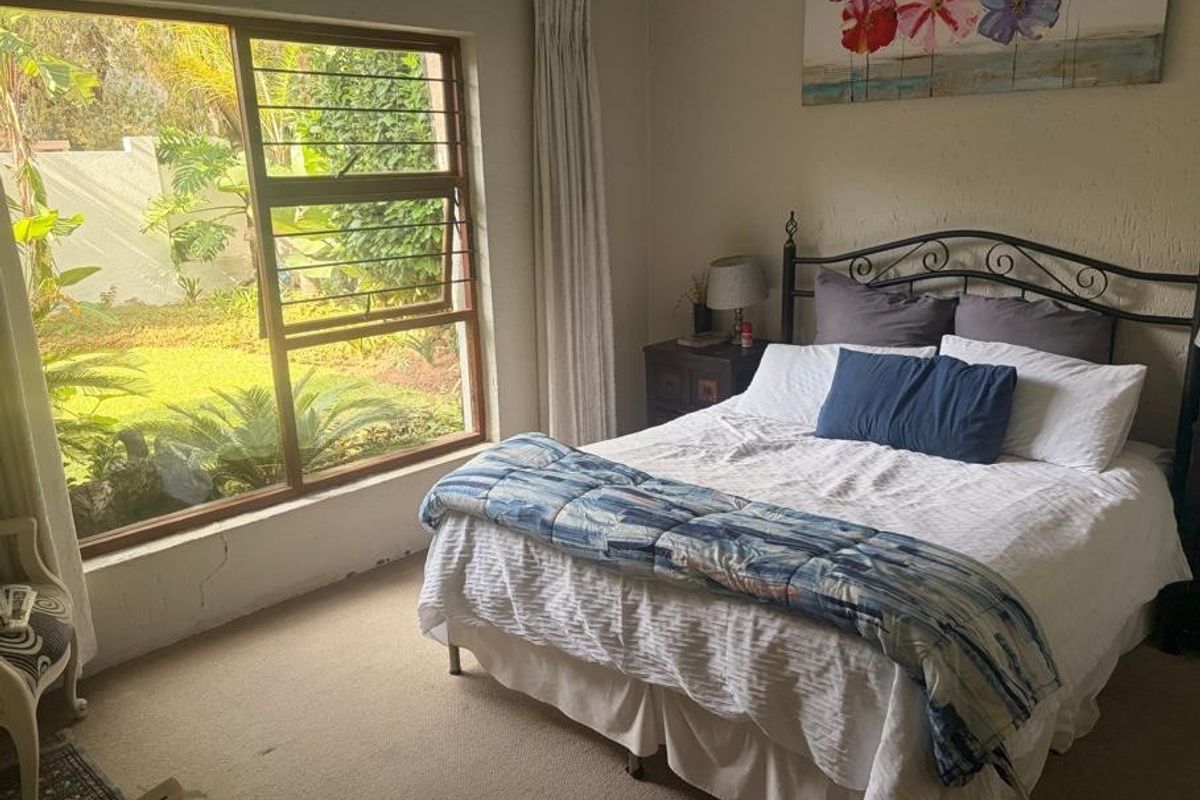 A bedroom featuring a large window with security bars, overlooking a garden, with carpeted flooring and visible wall cracks below the window frame.