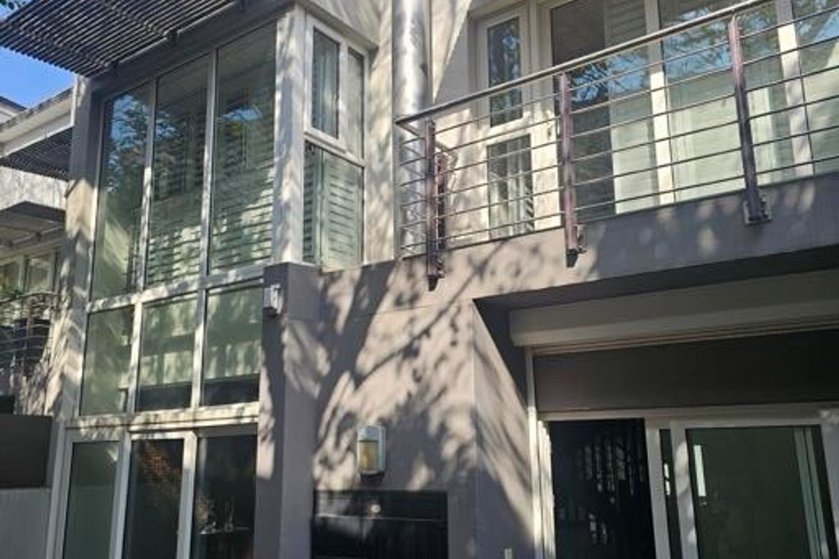Rear elevation of a modern townhouse featuring a built-in braai, balcony with stainless steel railing, large sliding glass doors, and metal sun louvres.