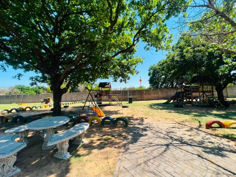 A spacious communal garden area featuring mature trees, paved pathways, a concrete boundary wall, and fixed outdoor seating, suitable for recreational use.