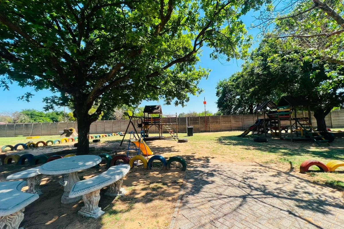 A spacious communal garden area featuring mature trees, paved pathways, a concrete boundary wall, and fixed outdoor seating, suitable for recreational use.