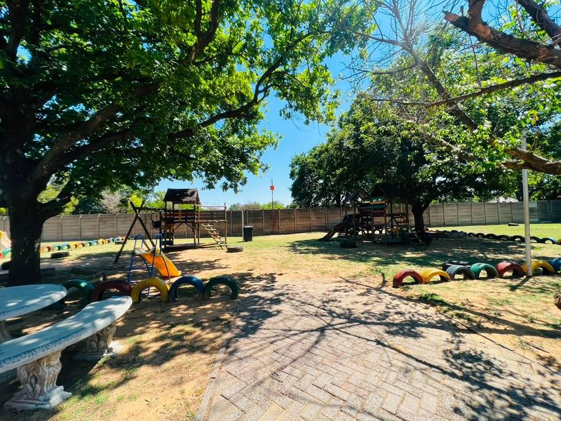 A spacious communal garden area featuring mature trees, paved pathways, concrete seating, and established play equipment within a secure fenced perimeter.