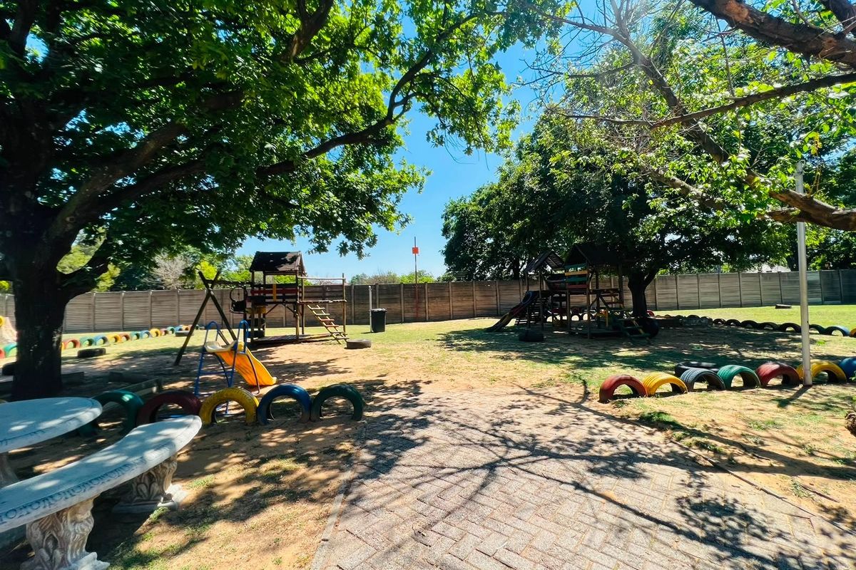 A spacious communal garden area featuring mature trees, paved pathways, concrete seating, and established play equipment within a secure fenced perimeter.
