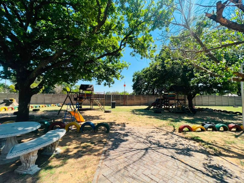 A spacious communal garden area featuring mature trees, a paved walkway, and established playground equipment enclosed by a concrete boundary wall.