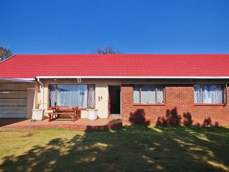 A single-story brick house featuring a prominent red metal roof, a front lawn, and an attached carport with a garage door.