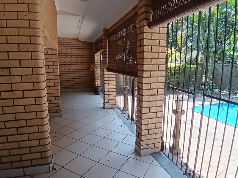 A covered patio area featuring exposed brick pillars and tiled flooring, overlooking a swimming pool and garden area.