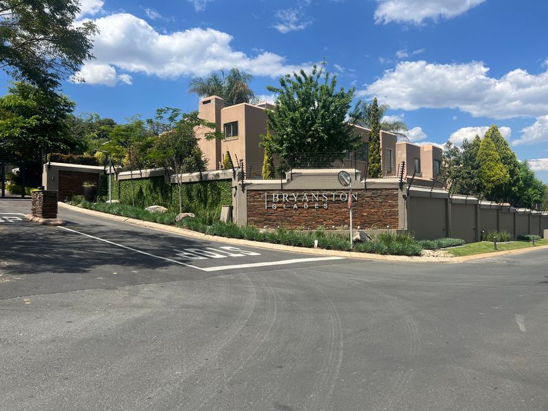Street view of the Bryanston Glades residential complex entrance, featuring stone-clad signage, secure perimeter walling with electric fencing, and well-maintained landscaping.