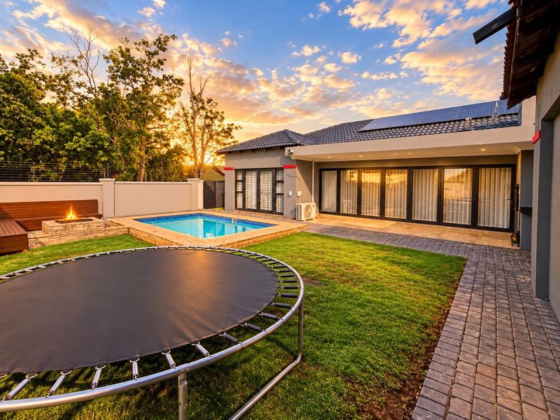 A beautifully landscaped rear garden featuring a swimming pool, built-in fire pit, paved pathways, and modern home exterior with solar panels at sunset.