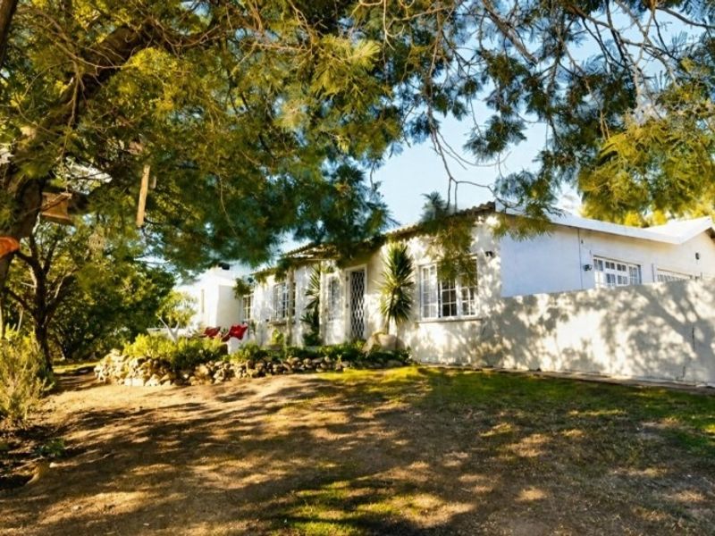 A charming farm-style residence featuring a white facade, tiled roof, and established garden surroundings with mature trees.