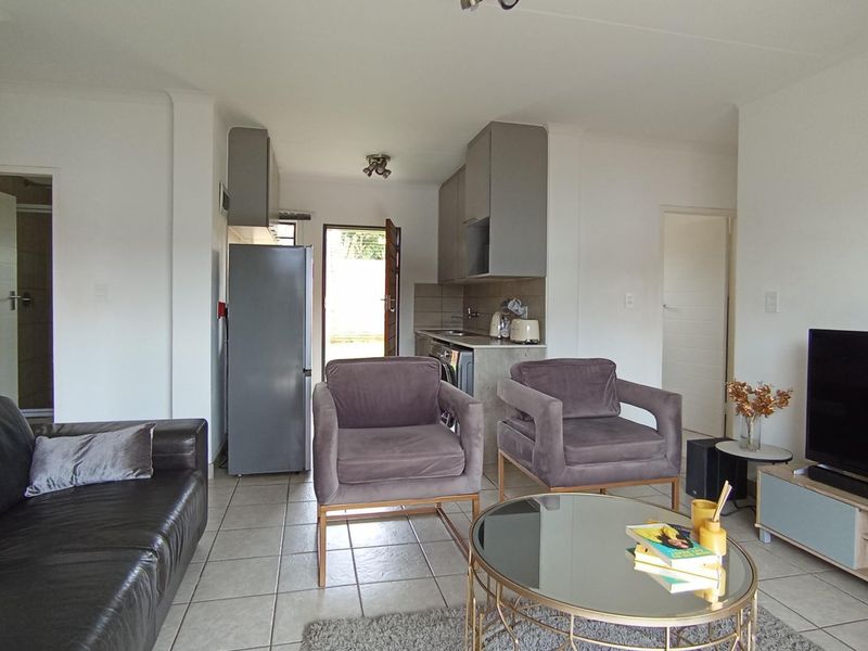 Open-plan living area featuring tiled flooring and a compact kitchen with built-in cabinetry and countertop space, leading to an exterior entrance.