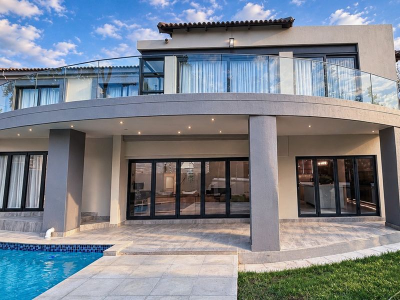 A modern rear elevation featuring a spacious tiled patio, swimming pool, and a curved upper balcony with a sleek glass balustrade.