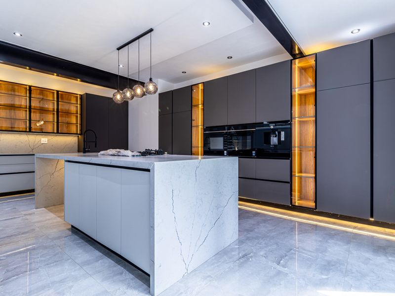 A modern, high-end kitchen featuring a large stone-topped island, sleek dark cabinetry, integrated appliances, and sophisticated ambient lighting throughout.