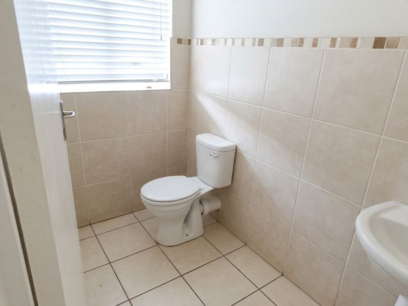 A functional guest bathroom featuring neutral ceramic wall and floor tiles, a standard toilet, and a wall-mounted basin with natural light from the window.