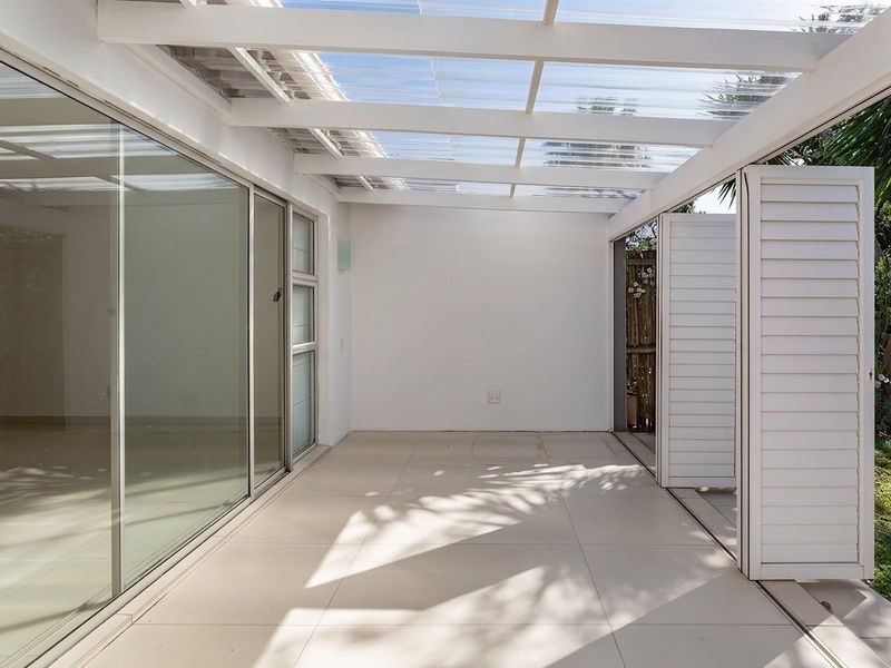 A bright, covered patio featuring tiled flooring, a translucent roof, and sliding glass doors, offering a seamless transition to the garden area.