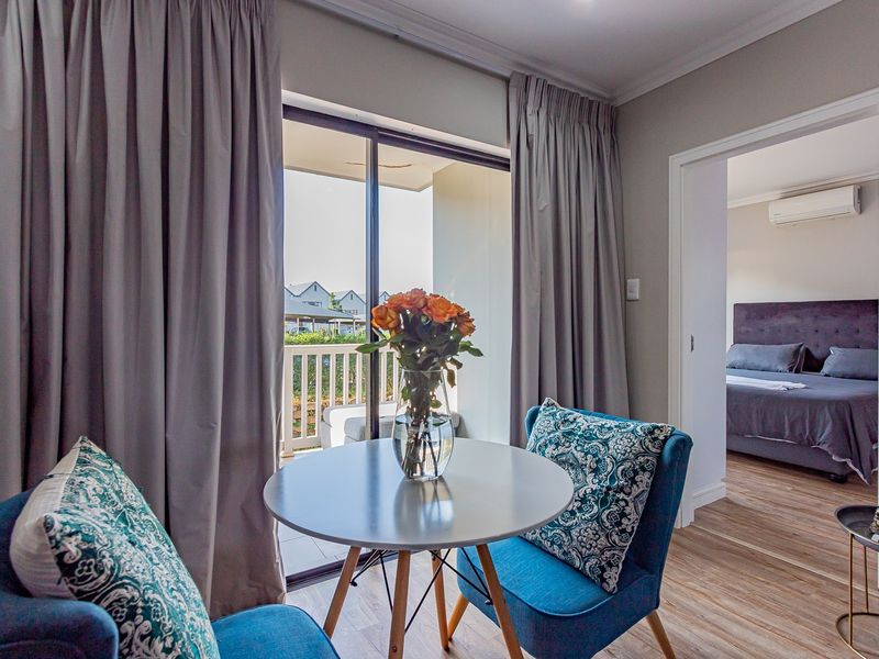 A bright living area featuring laminate flooring, coving, and a sliding glass door leading to a balcony, with a view into the adjacent bedroom.