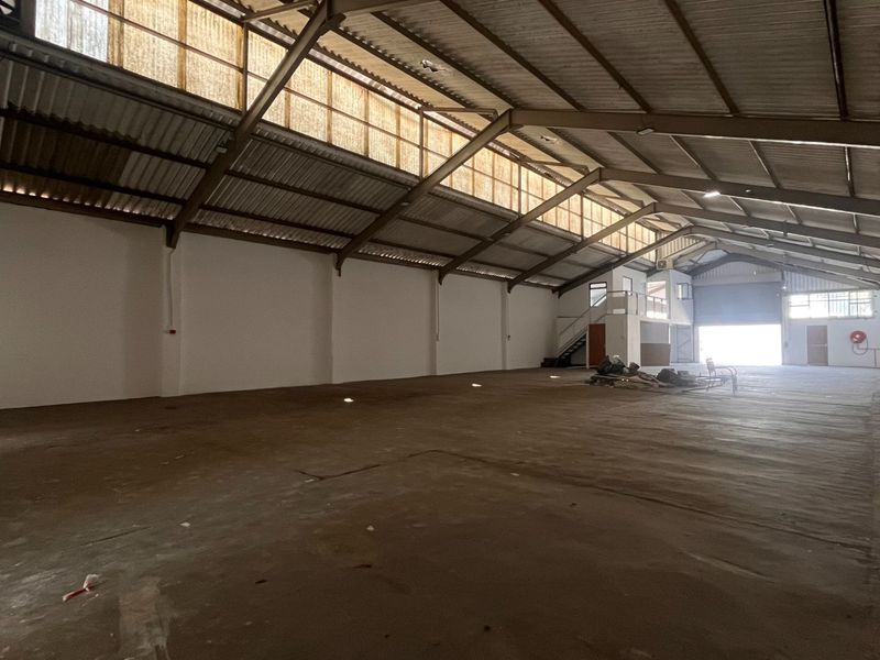 Spacious industrial warehouse featuring high ceilings with exposed steel trusses, clerestory windows for natural light, concrete flooring, and a roller shutter door.