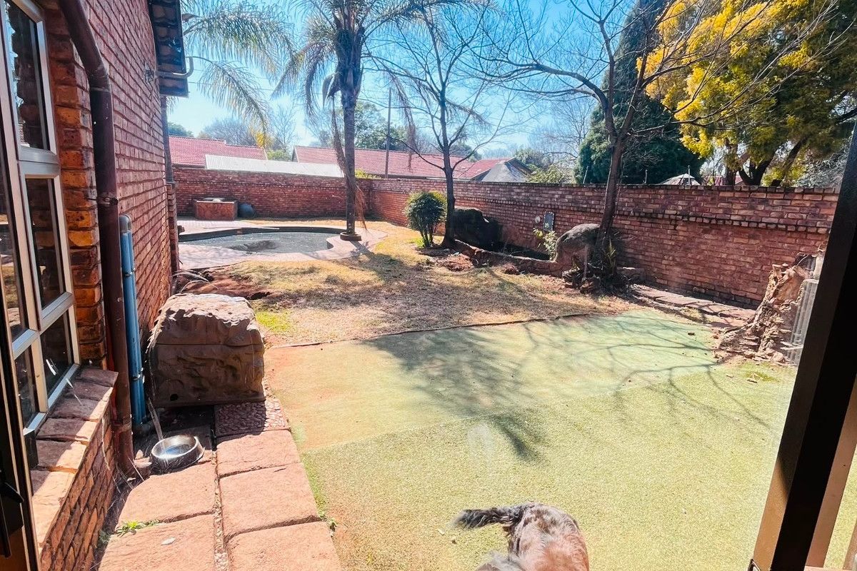 Rear garden area featuring a swimming pool, artificial lawn, and brick boundary walls, viewed from under a thatched roof patio.