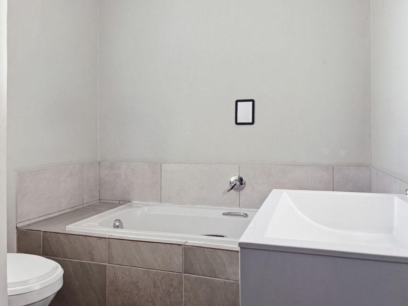 A clean, modern bathroom featuring a built-in bathtub with wall-mounted mixer, a vanity basin, and a toilet with neutral tiled finishes.