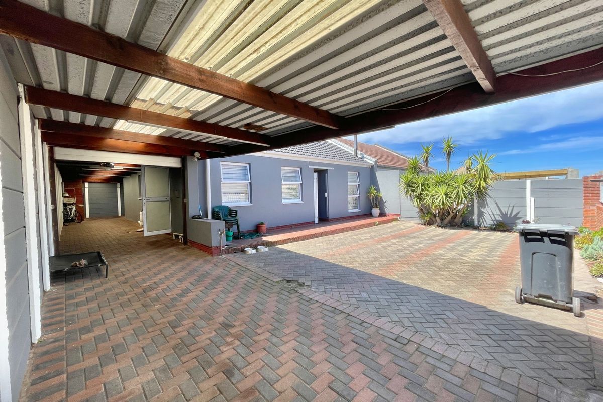 A spacious rear exterior area featuring a covered patio with timber beams, paved driveway, and a secure boundary wall.