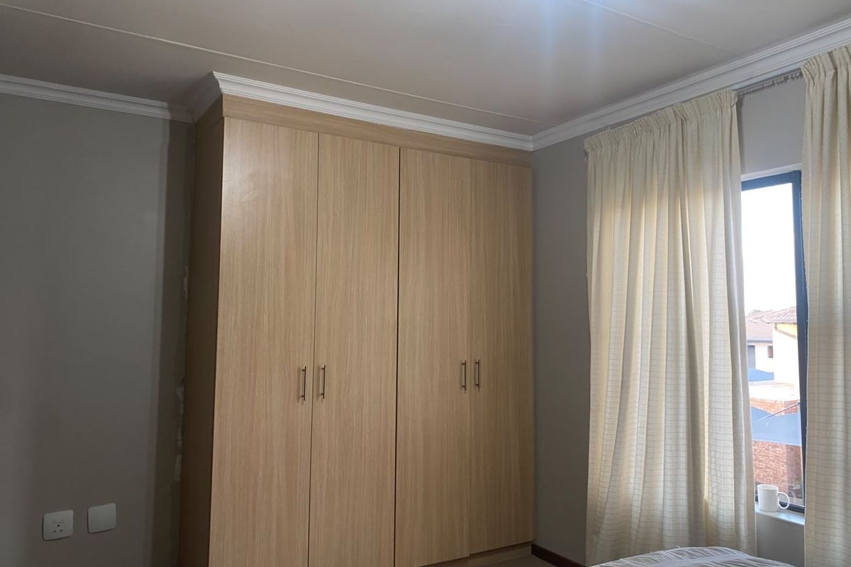 A well-maintained bedroom featuring light wood-finish built-in wardrobes, crown molding, and a window with curtains.