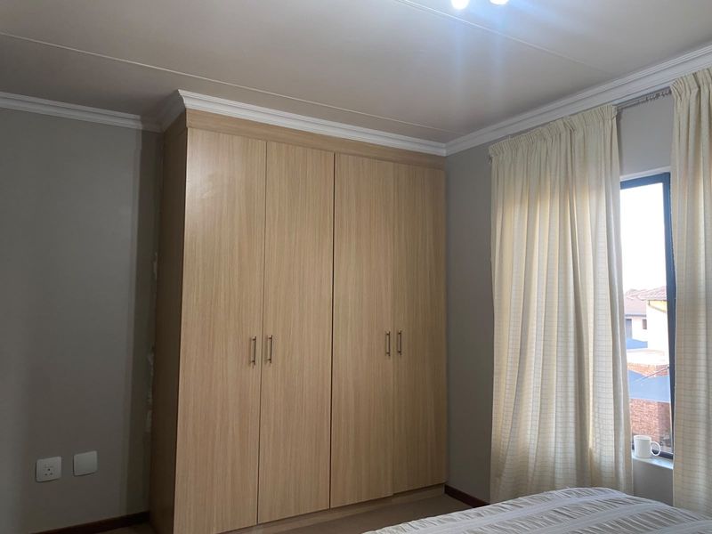 A well-maintained bedroom featuring light wood-finish built-in wardrobes, crown molding, and a window with curtains.