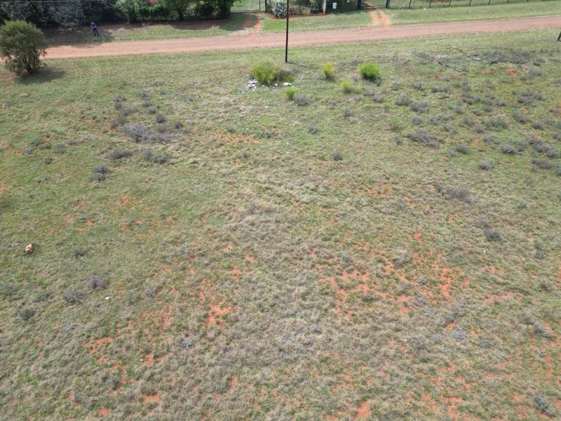 An aerial view of a vacant residential plot featuring natural grass cover, perimeter fencing, and adjacent dirt road access.