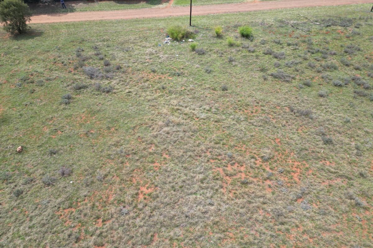 An aerial view of a vacant residential plot featuring natural grass cover, perimeter fencing, and adjacent dirt road access.