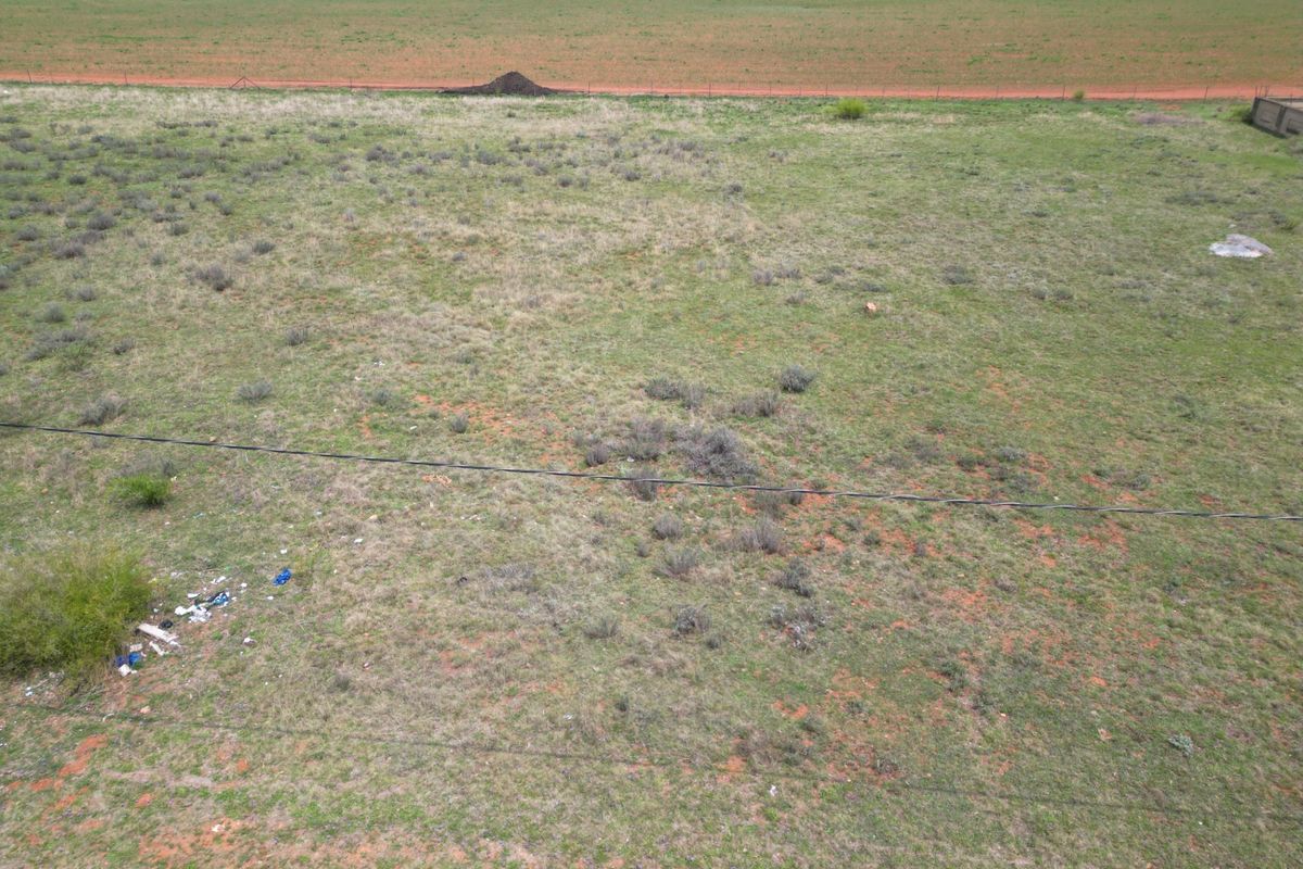 An aerial view of a vacant residential plot featuring natural grass cover, perimeter fencing, and overhead power lines.