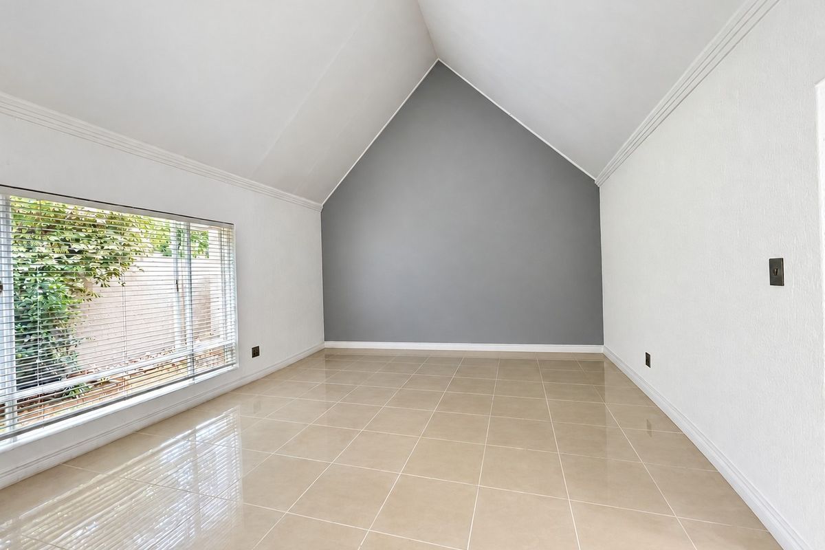A bright, empty bedroom featuring a high vaulted ceiling, neutral tiled flooring, and a large window with venetian blinds.
