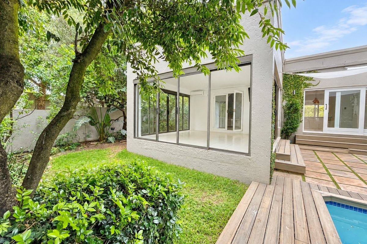 Rear exterior view featuring a modern glass-enclosed living space, wooden decking, swimming pool, and a well-maintained garden area.