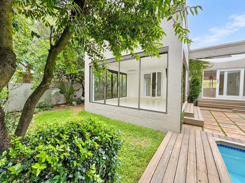 Rear exterior view featuring a modern glass-enclosed living space, wooden decking, swimming pool, and a well-maintained garden area.