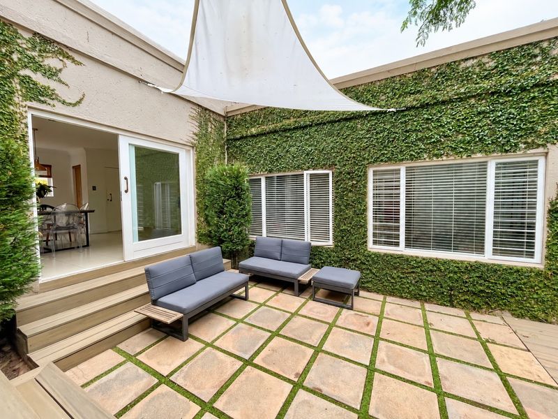 A charming paved courtyard patio featuring a shade sail, climbing ivy walls, and direct access to the interior through a sliding glass door.