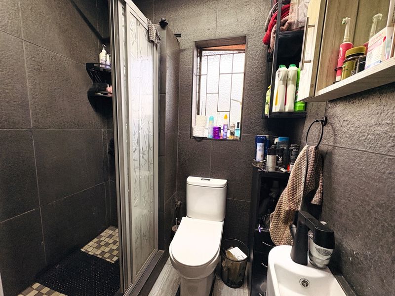 Compact bathroom featuring dark tiled walls, a shower enclosure, a toilet, and a wall-mounted basin with security bars on the window.