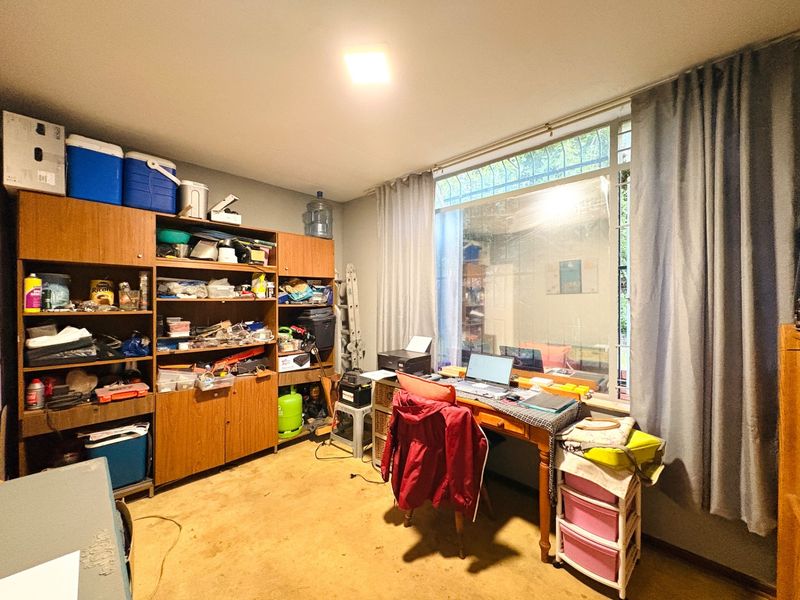 A functional office space featuring built-in cabinetry, a large window with security bars, and neutral wall finishes, offering potential for a dedicated workspace.