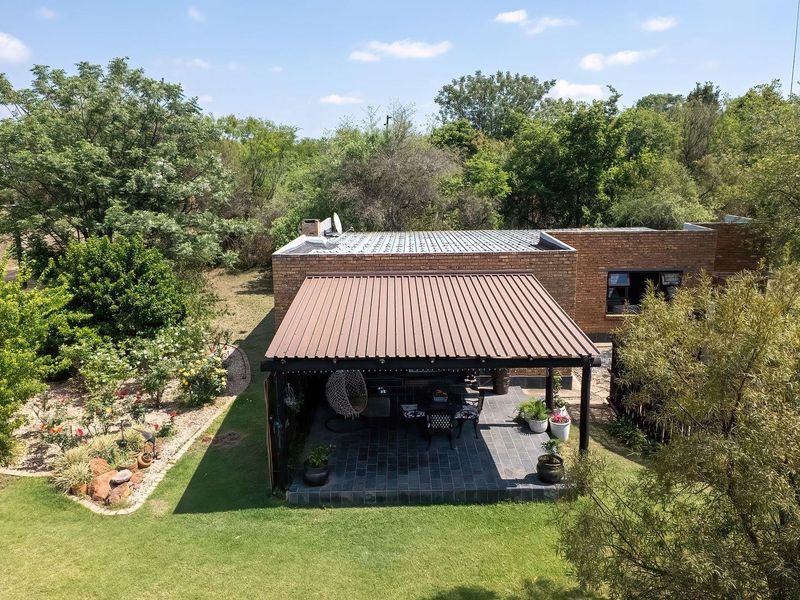 Aerial view of a brick farmhouse featuring a covered patio area with tiled flooring and a corrugated metal roof, surrounded by established garden landscaping.
