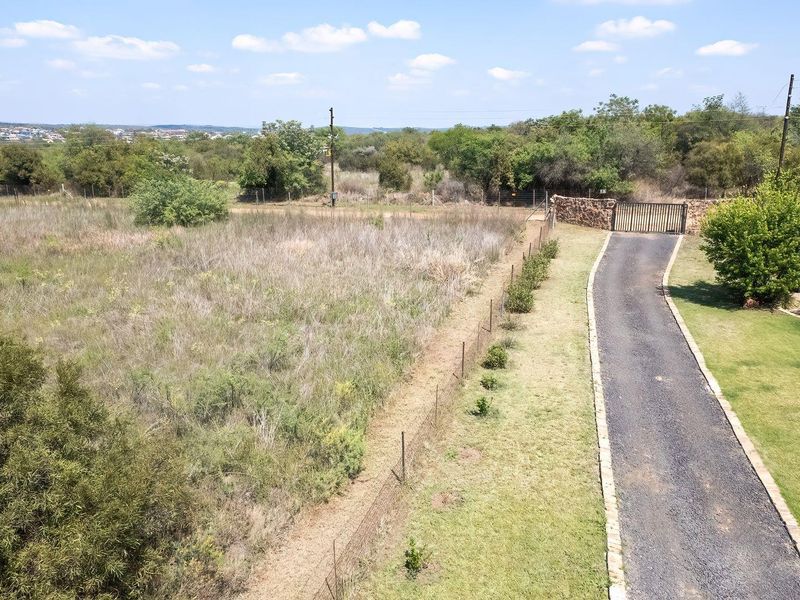 Aerial view of the property entrance featuring a paved driveway, stone boundary wall, metal gate, and expansive surrounding landscape.