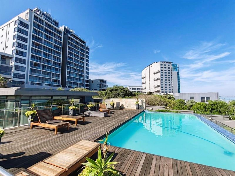 A well-maintained outdoor swimming pool area featuring timber decking and a modern glass balustrade, set within a high-rise apartment complex.