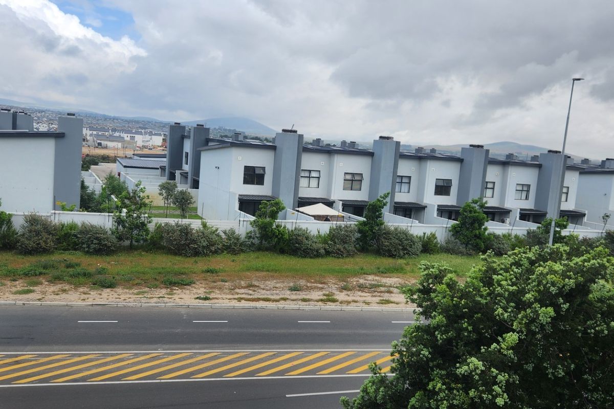 A view of the rear elevation of a modern apartment complex, featuring uniform architectural design, boundary walls, and proximity to a public road.