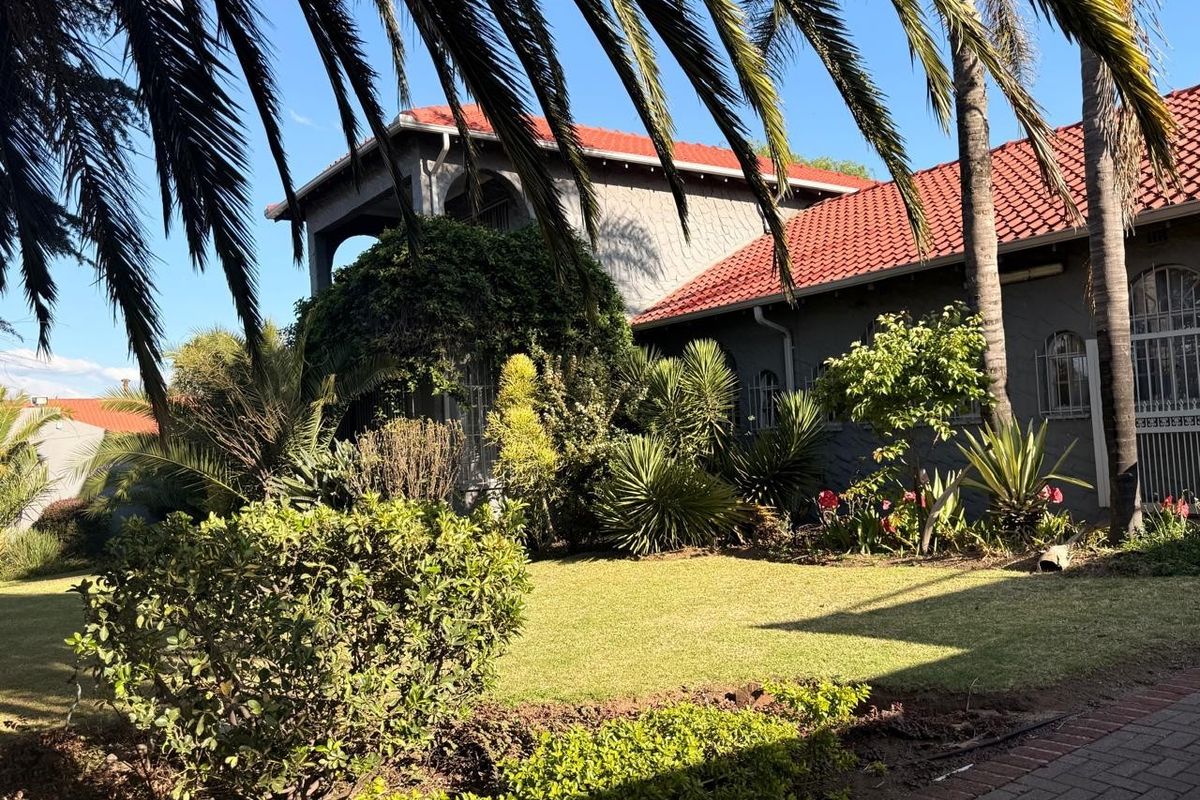 A well-maintained rear garden featuring a lush lawn, mature palm trees, and a house exterior with terracotta roof tiles and grey rendered walls.