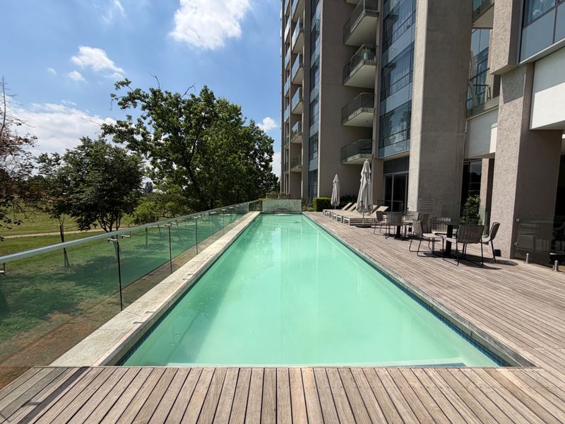 A modern lap pool area featuring timber decking, a glass balustrade, and views of the surrounding landscape, set against a contemporary apartment building.