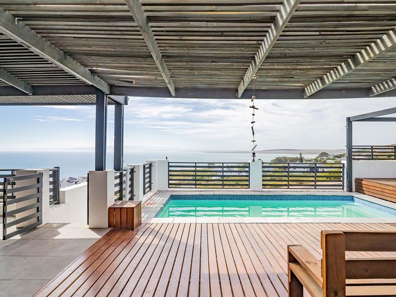 An elevated outdoor pool area featuring timber decking, a pergola, and a built-in braai, offering expansive coastal views.