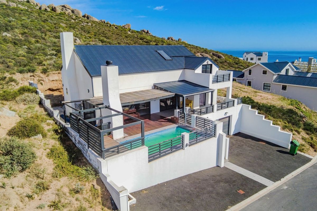 Aerial view of a modern coastal house featuring a swimming pool, timber deck, pergola, and balcony, set against a hillside landscape.
