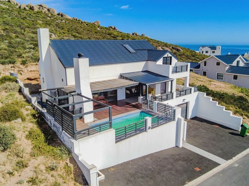 Aerial view of a modern coastal house featuring a swimming pool, timber deck, pergola, and balcony, set against a hillside landscape.