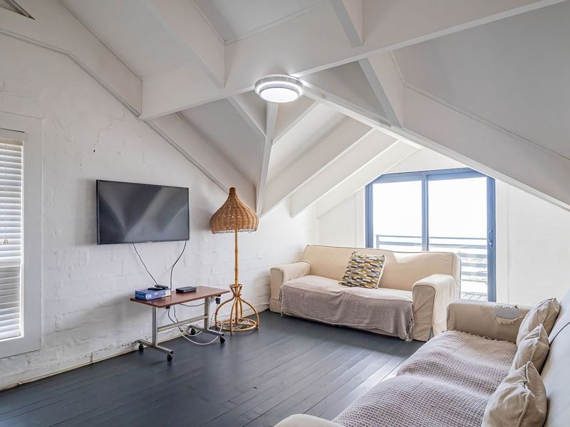 A bright living room featuring vaulted ceilings with exposed rafters, painted brick walls, dark timber flooring, and access to an outdoor balcony.